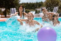 Ein blonder Junge, ein braunhaariges Mädchen mit Zöpfen und ihre Eltern spielen in einem Freibad mit einem lilanen Ball