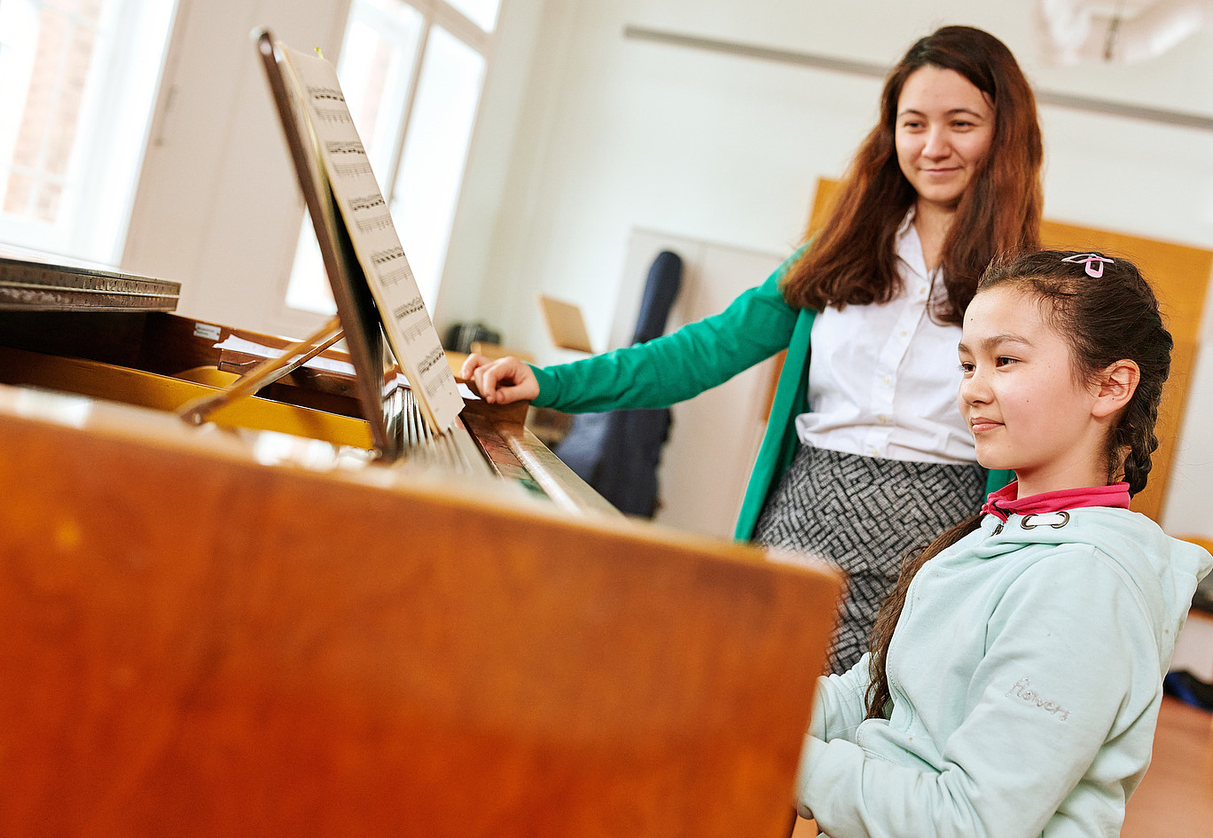 Ein junges Mädchen spielt etwas auf einem Klavier und eine Frau steht daneben, schaut auf das Klavier und lehnt an dem Klavier.