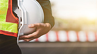 Hand hält Helm an menschlichen Oberkörper vor unscharfer Bauzaunkulisse