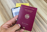Eine Hand hält einen Reisepass. Im Hintergrund sind unscharf ein Handy und ein Impfpass zu sehen.