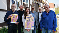 Gruppenfoto mit (von links) Bernd Groeger und Joachim Kleinmanns vom Heimatverein, Nora Moos (Ehrenamtskoordinatorin der Stadt Detmold), Werner Zahn (Heimatverein), Ortsbürgermeister Rainer Friedrich und Heinz Heerde, ebenfalls vom Heiligenkirchener Heimatverein. Sie halten Plakate vom Ortsteilfest in die Kamera.