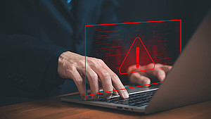 Hände, die auf einer Laptop-Tastatur tippen und darüber ein Warnzeichen und unscharfer Programmier-Code in rot