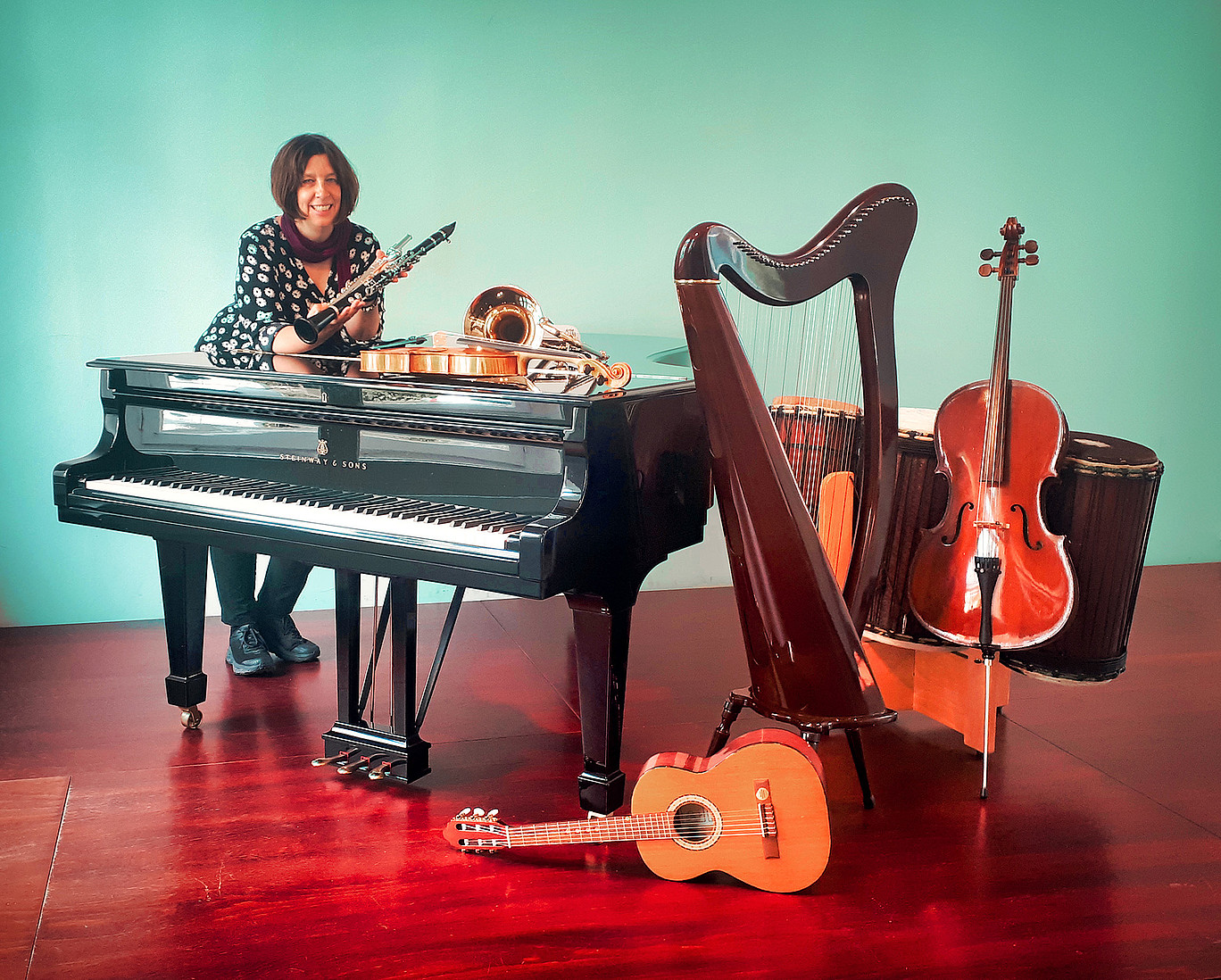 Eine Frau lehnt an einem Klavier mit einer Querflöte und einer Klarinette in den Händen. Auf, an und neben dem Klavier sind weitere Instrumente zu sehen.