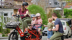 Eine Mutter sitzt mit ihren zwei Töchtern auf einem Fahrrad. Der Vater hockt mit einem weiteren Kind direkt neben dem stehenden Fahrrad.