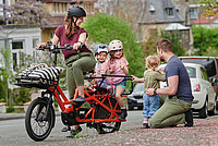 Eine Mutter sitzt mit ihren zwei Töchtern auf einem Fahrrad. Der Vater hockt mit einem weiteren Kind direkt neben dem stehenden Fahrrad.