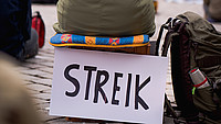 Ein Streik-Schild lehnt an einem Wasserkasten, auf welchem eine Person sitzt.
