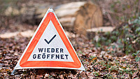 Warndreieck auf Waldboden mit der Aufschrift "WIEDER GEÖFFNET" und einem Häkchensymbol darüber