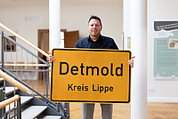 Bürgermeister Frank Hilker steht in der zweiten Etage des Rathauses der Stadt Detmold und hält ein gelbes Ortsschild von Detmold in die Höhe