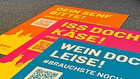 Drei A1-Neonplakate auf einem Teppichboden mit den Aufschriften "DEIN SENF BITTE!", "ISS DOCH KÄSE!" und "WEIN DOCH LEISE!"