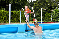 Ein Vater und ein Kind mit Schwimmflügeln steigen gemeinsam über eine Leiter aus dem Becken eines Freibades