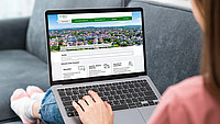 Eine Frau (im Anschnitt und unscharf) sitzt auf einem grauen Sofa und hat einen aufgeklappten Laptop auf den Beinen stehen, auf dem Bildschirm ist die Startseite des Serviceportals der Stadt Detmold zu sehen.