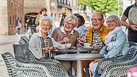 Eine Gruppe von vier Rentnerinnen und Rentnern sitzt draußen an einem Tisch in der Detmolder Innenstadt und schaut sich gemeinsam lachend etwas an