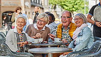 Eine Gruppe von vier Rentnerinnen und Rentnern sitzt draußen an einem Tisch in der Detmolder Innenstadt und schaut sich gemeinsam lachend etwas an