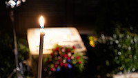 Im Vordergrund ist scharf eine brennende Kerze zu sehen, im Hintergrund ist unscharf ein Gedankstein zu sehen