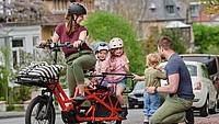 Eine lächelnde Frau sitzt mit zwei kleinen Kindern auf einem speziellen Fahrrad. Davor kniet ein Mann mit einem Kleinkind.