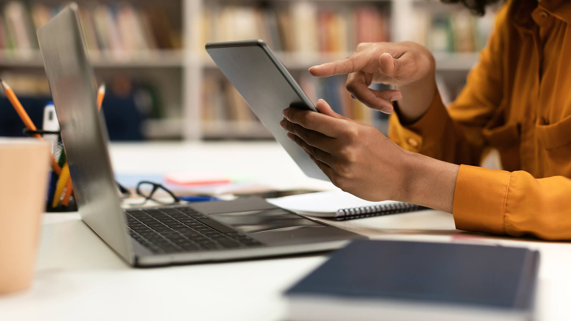 Eine Person zeigt auf etwas auf einem Tablet. Hinter dem Tablet ist im Anschnitt ein Laptop zu sehen und im Hintergrund sind unscharfe Bücherregale abgebildet.