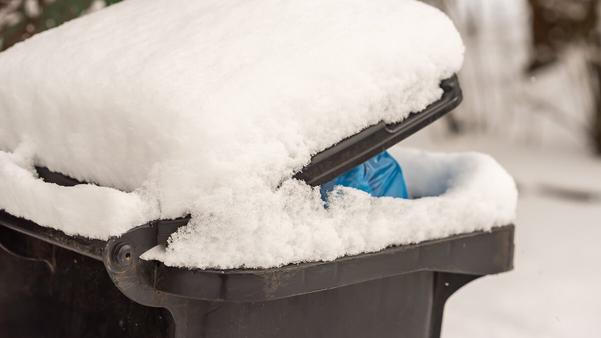 Seitenaufnahme einer grauen Mülltonne, welche mit einer Schicht Schnee bedeckt ist