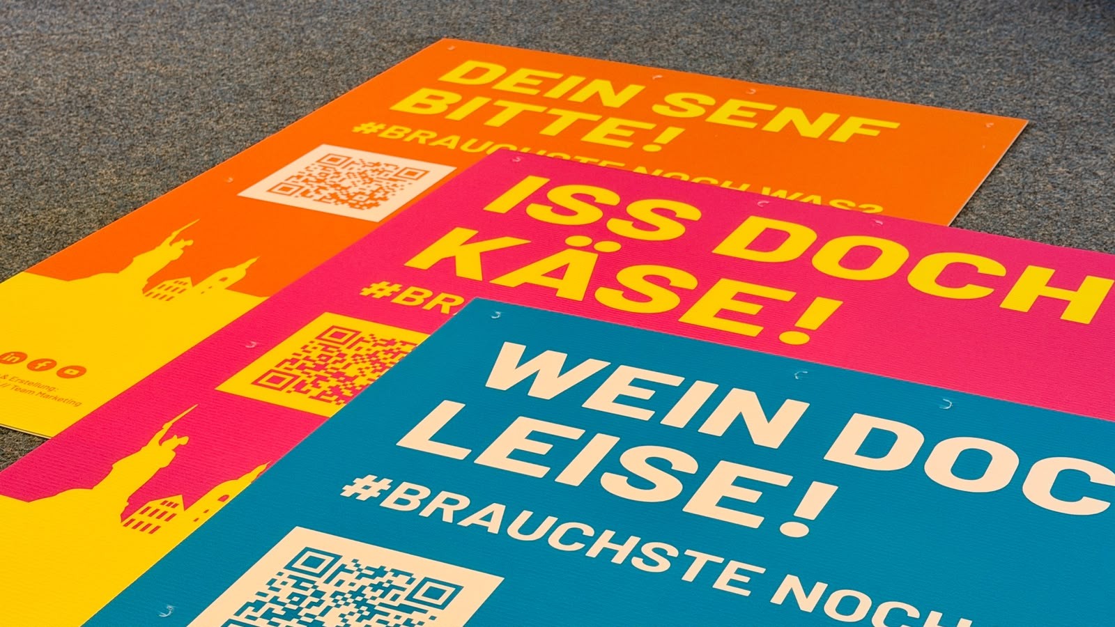 Drei A1-Neonplakate auf einem Teppichboden mit den Aufschriften "DEIN SENF BITTE!", "ISS DOCH KÄSE!" und "WEIN DOCH LEISE!"